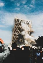 An office building collapses in the background while, in the foreground, several people take photos of the catastrophe on their phone